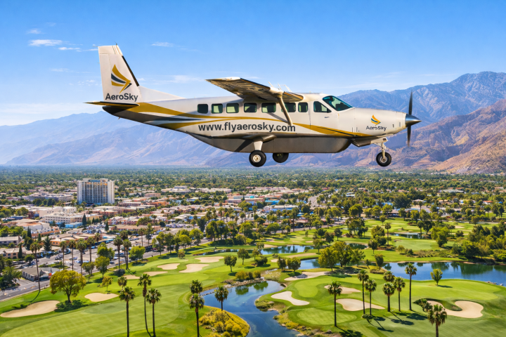 Small airplane flying over a cityscape with mountains and golf course below.