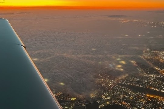 Wing of a plane flying over a cityscape at sunset with visible city lights.